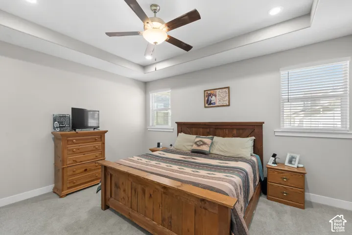 Tray ceiling with recessed lighting, ceiling fan, two windows with blinds, and light gray wall paint