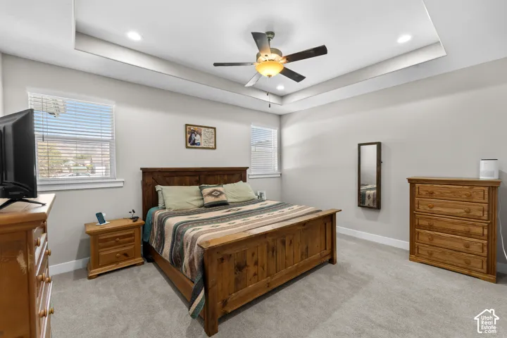Tray ceiling with recessed lighting, ceiling fan with integrated light, two windows with blinds, and light gray carpet flooring