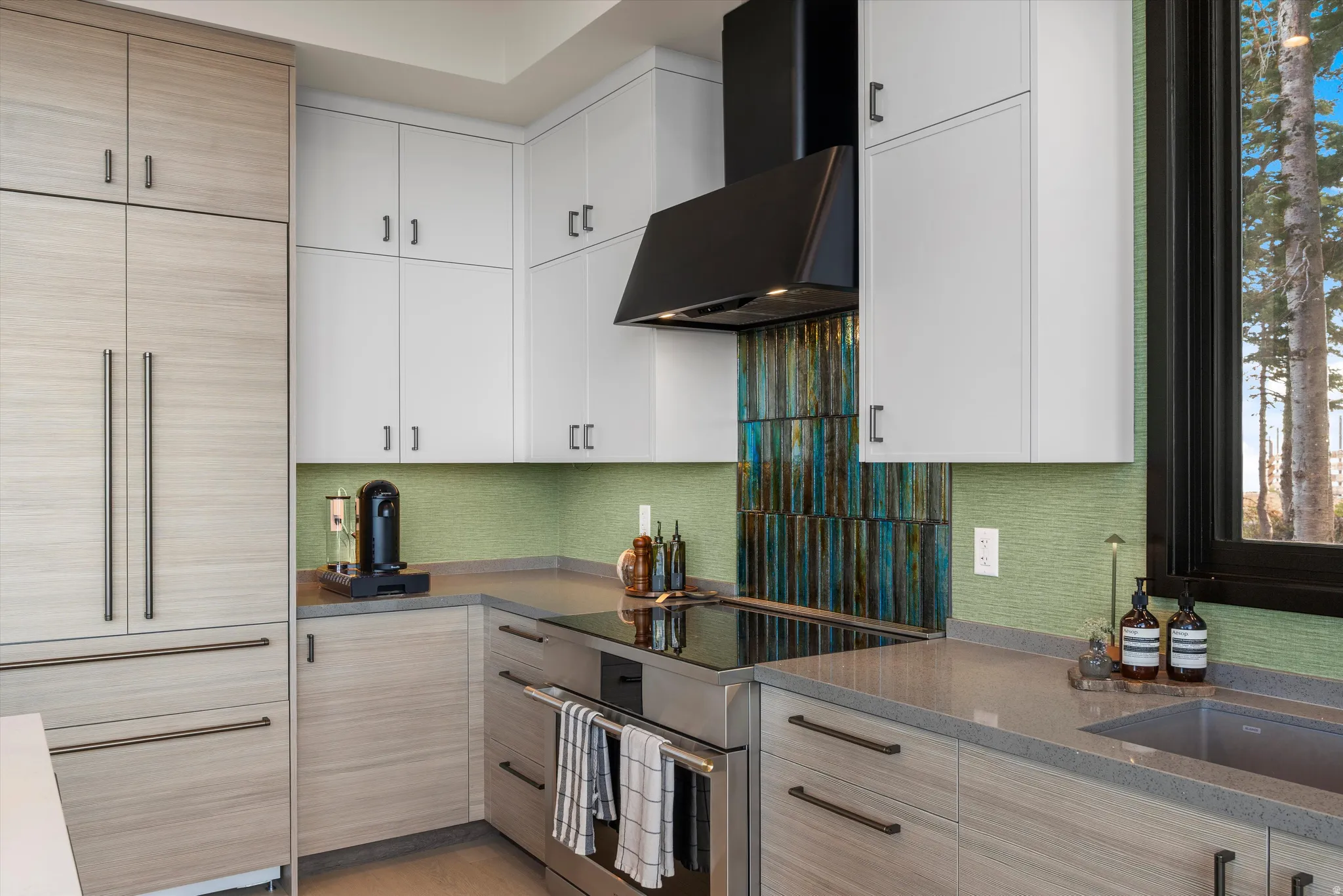 Integrated refrigerator, stainless steel oven, cooktop, and range hood, light wood-finish lower cabinetry, white upper cabinetry, and a textured green and blue tile backsplash