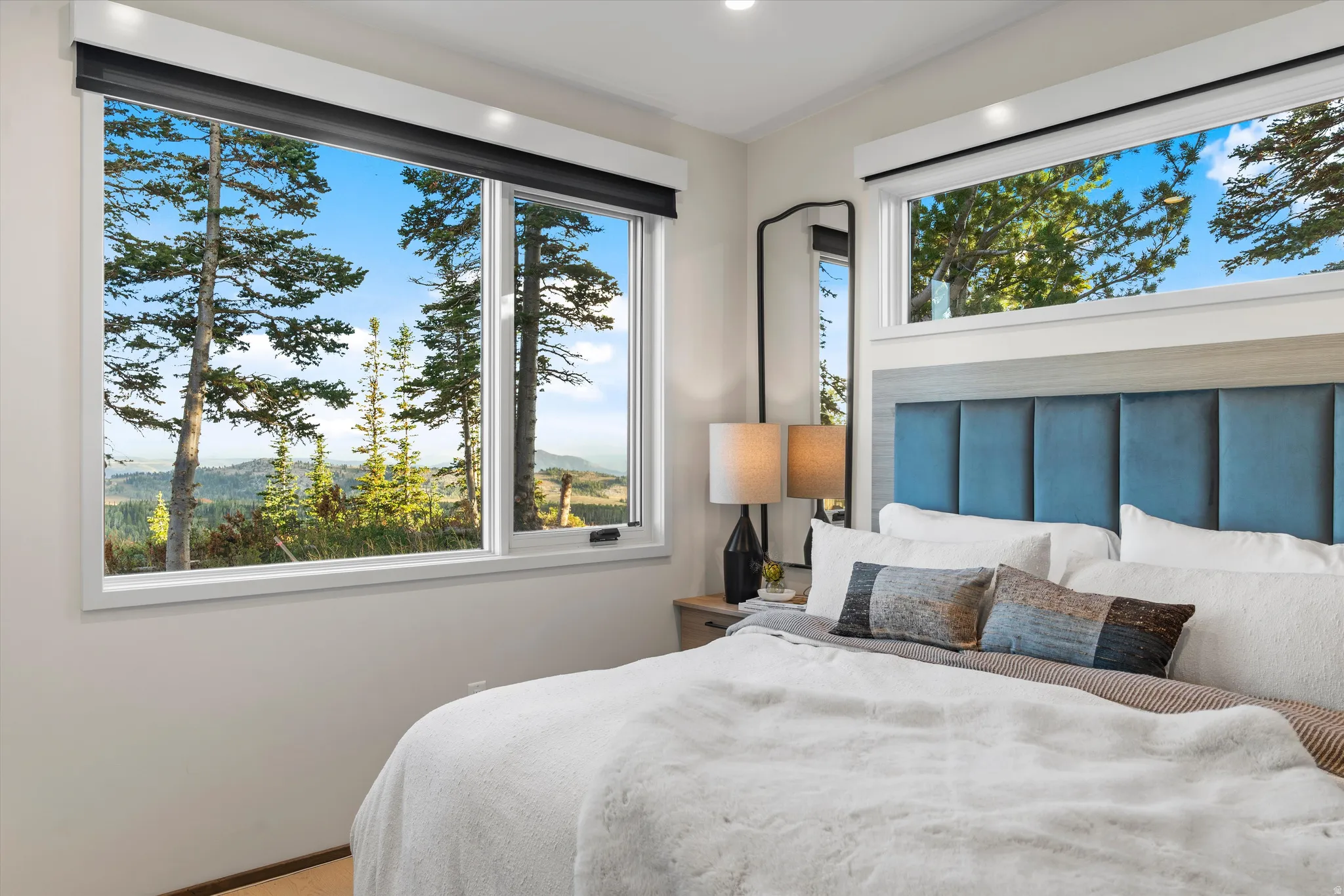 Primary bedroom featuring expansive multi-pane windows framing mountain and forest scenery, a blue upholstered headboard, hardwood flooring, recessed ceiling lighting, and a transom window above for enhanced natural light