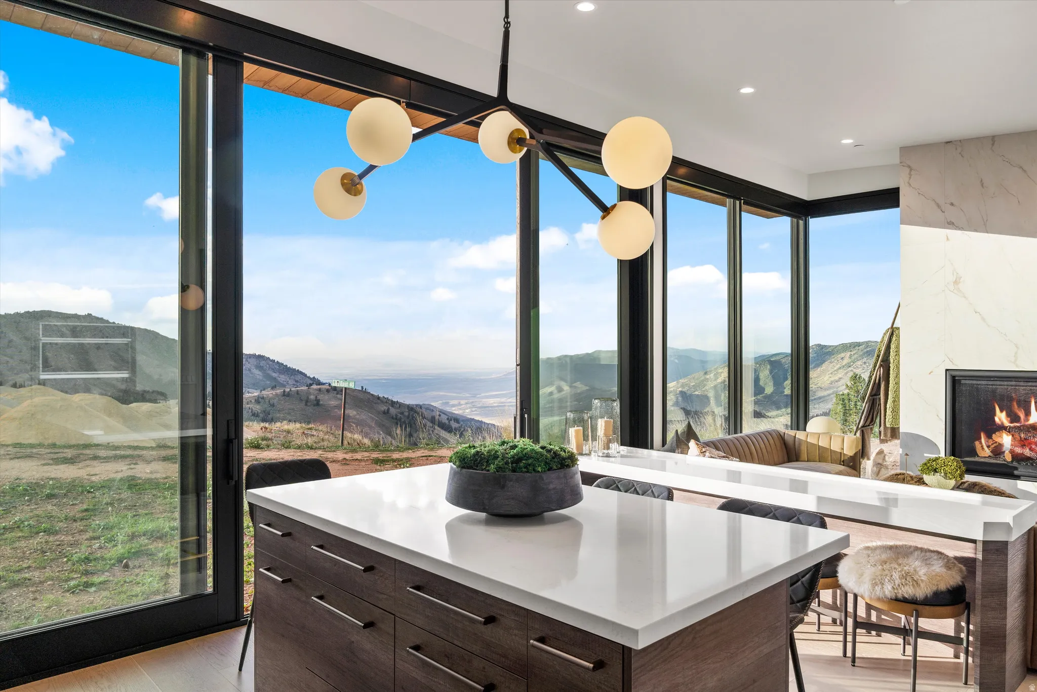 Expansive sliding glass doors, contemporary fireplace with large format tile surround, wood-finish cabinetry, white countertop surfaces, and modern spherical chandelier