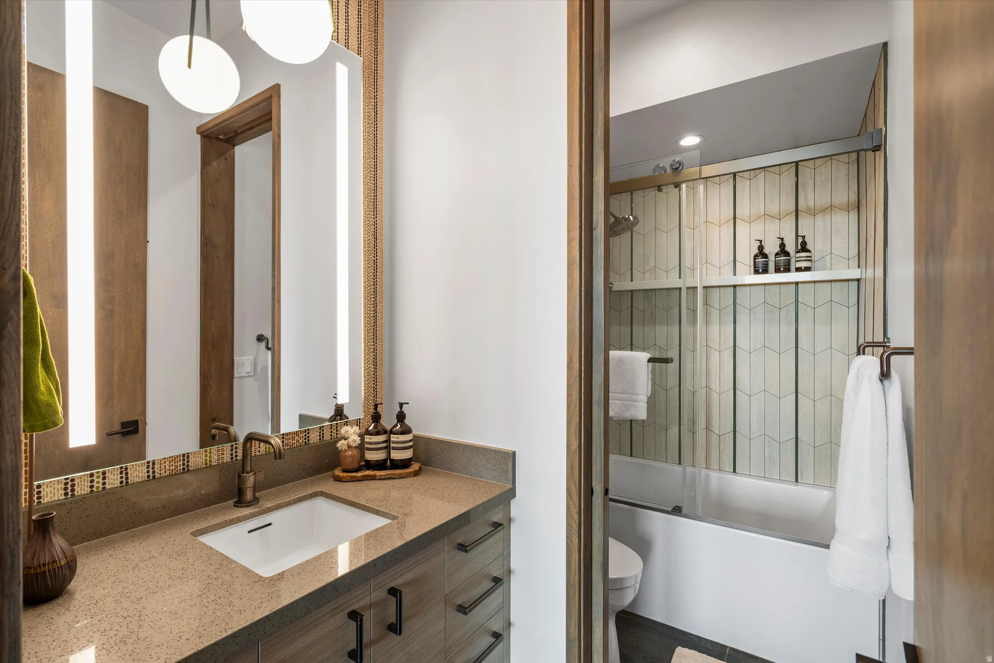 Vanity with integrated sink and bronze-finish faucet, illuminated mirror with accent lighting, and a bathtub with a tiled surround