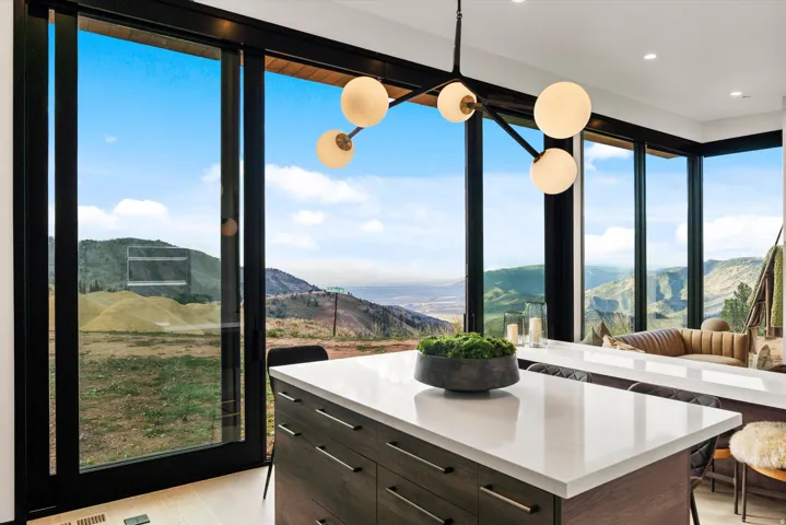 Expansive window wall with sliding glass door, contemporary light fixture, large kitchen island with light-toned countertop, and dark wood-finish cabinetry