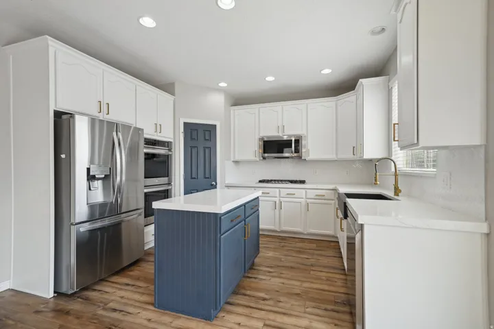 White cabinetry with gold-tone hardware, stainless steel appliances, wood-finish flooring, white countertop surfaces, and a blue island with beadboard paneling