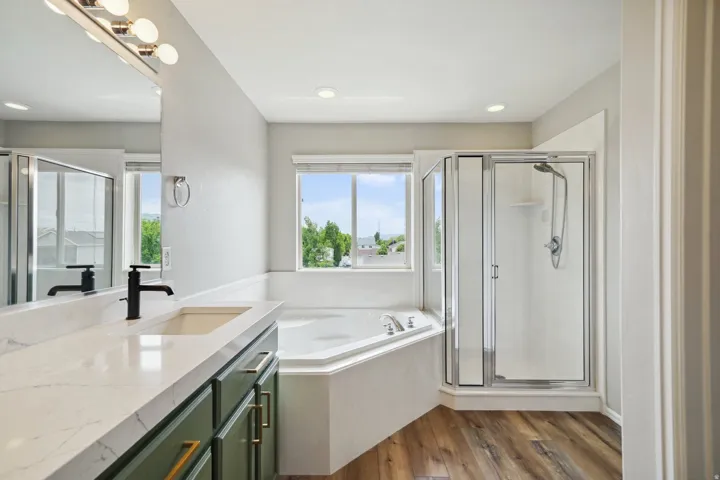 Wood-finish flooring, corner soaking tub, glass-enclosed shower, dual-sink vanity with dark green cabinetry, and white countertop