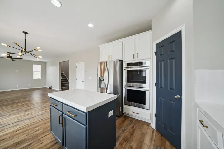 Kitchen island with a light-colored countertop and dark-toned cabinetry, stainless steel double ovens, white upper cabinetry with brass-toned hardware, wood-finish flooring, and a contemporary chandelier