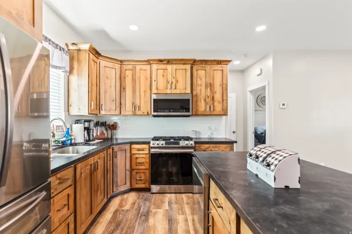 Kitchen featuring wood-finish cabinetry, stainless steel appliances, dark countertops, a tile backsplash, and wood-finish flooring