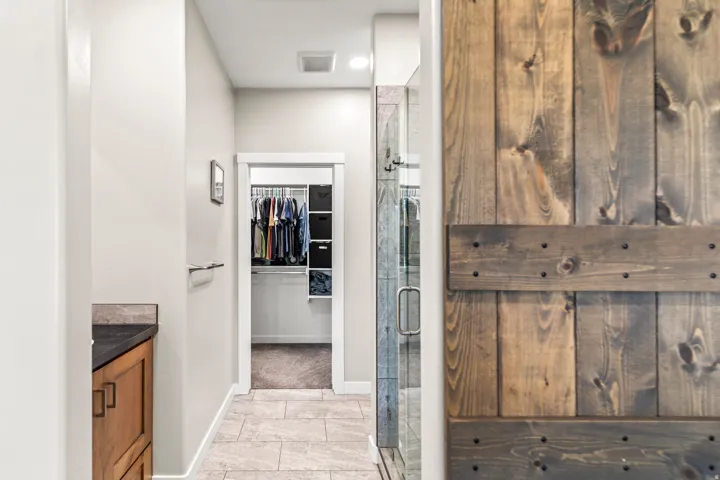 Wood-finish barn door, glass-enclosed shower, tiled flooring, wood vanity with dark countertop, and organized closet system