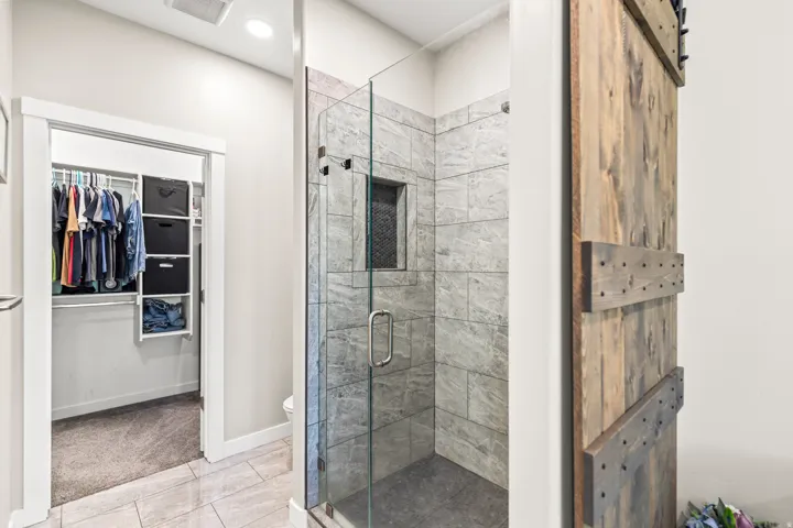 Primary bathroom featuring a frameless glass-enclosed shower with marble-look tile surround and flooring, polished chrome hardware, and direct access to a walk-in closet with built-in shelving and pegboard organization system