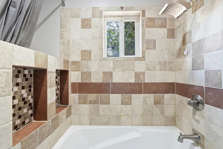 Bathtub with tiled surround, dual shower niches featuring mosaic tile accents, and a window for natural light