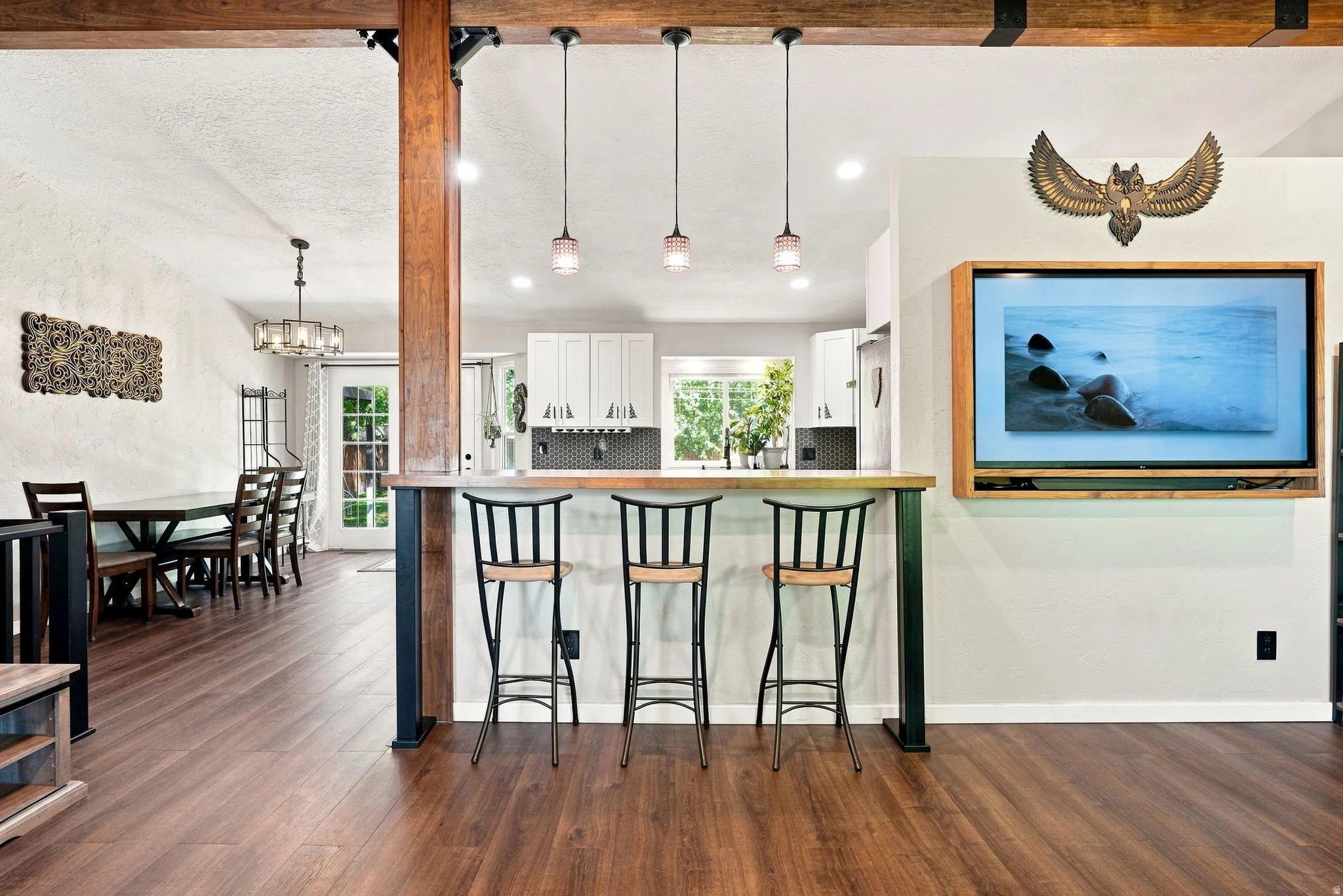 Wood-finish flooring throughout, exposed wood beams, breakfast bar with pendant lighting, white cabinetry, and a dark tile backsplash