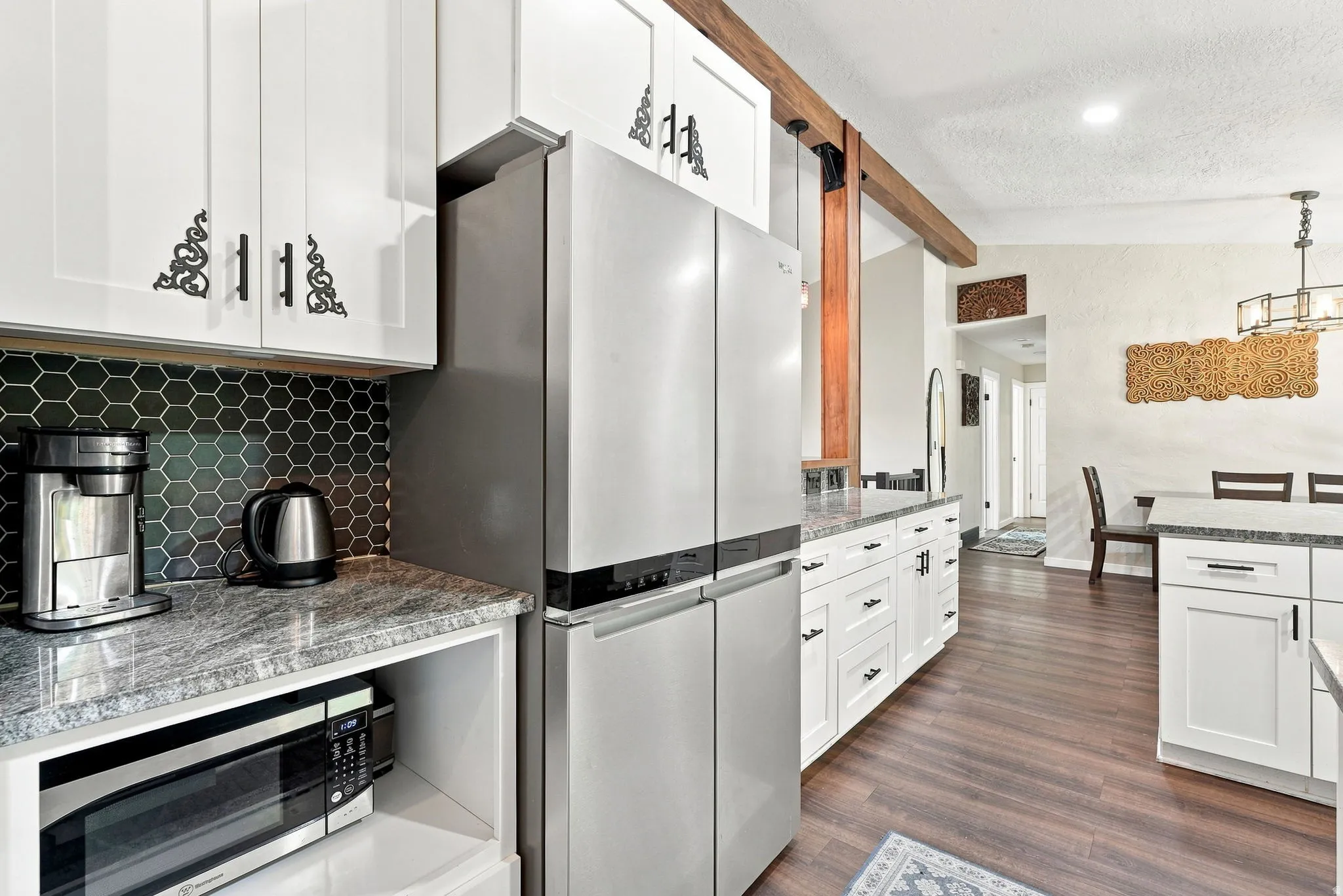 White cabinetry with decorative hardware, stainless steel refrigerator, granite countertops, dark hexagonal tile backsplash, and wood-finish flooring