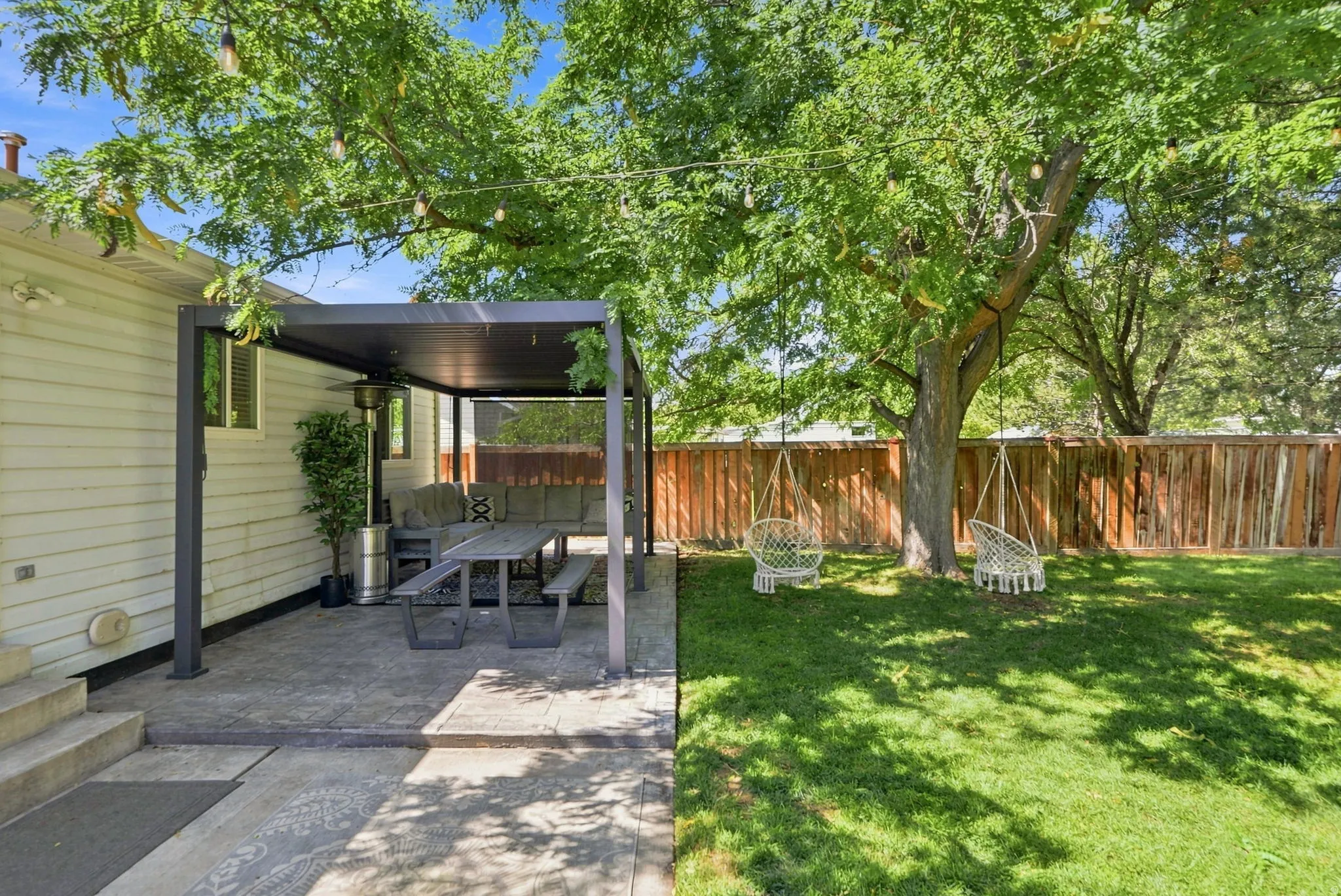 Expansive backyard featuring a shaded patio with a dark-frame pergola, concrete paver flooring, a manicured lawn, a mature tree, and a wooden privacy fence