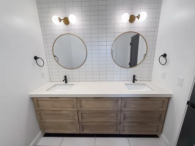 Dual vanity with wood-finish cabinetry, integrated sinks, white countertop, and individual circular mirrors