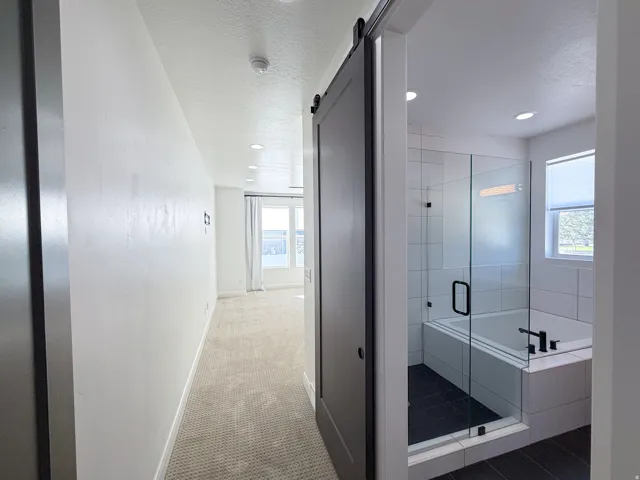 Sliding barn door entry to bathroom featuring a glass-enclosed shower, built-in bathtub, black fixtures, and a window