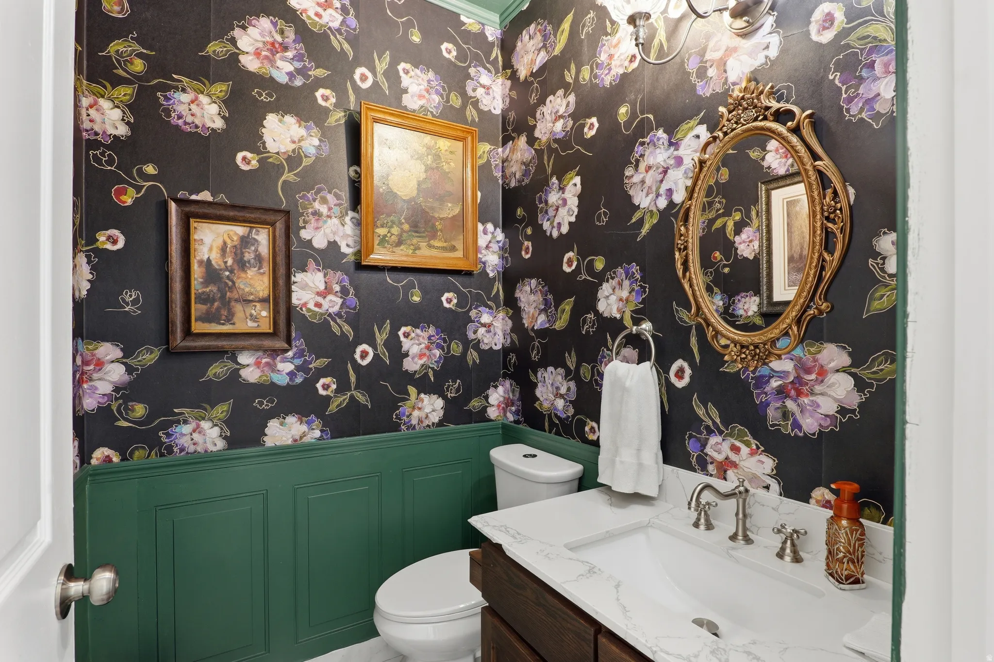 Elegant powder room featuring a single vanity with a white countertop and undermount sink, intricate floral wallpaper, green wainscoting, and a decorative wall mirror