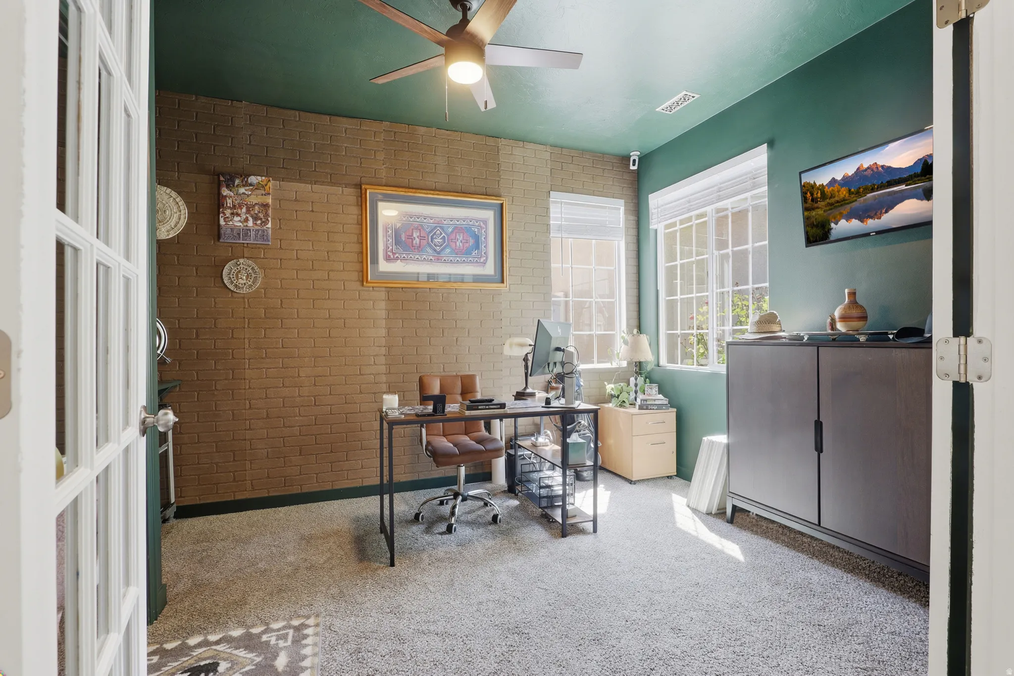 Carpeted room with a ceiling fan, two windows with blinds, a brick-textured accent wall, and a green painted wall