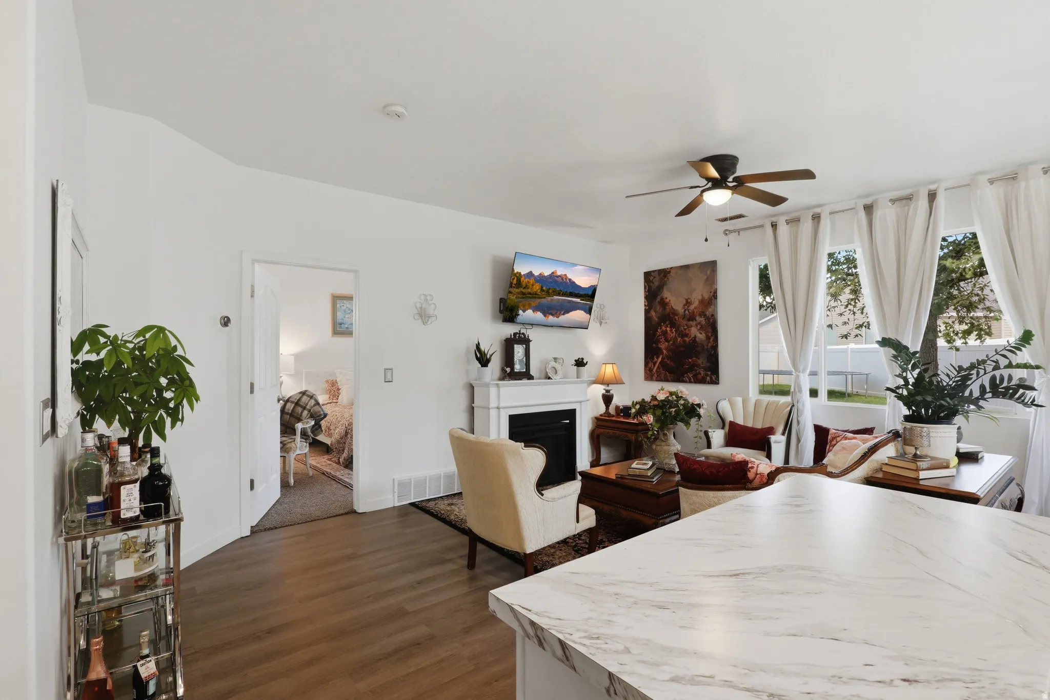 White countertop with subtle veining, wood-finish flooring, ceiling fan, white fireplace with mantel, and large windows