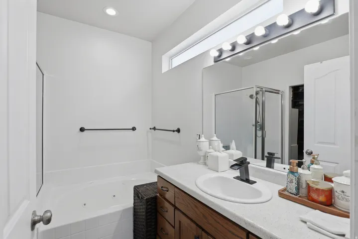 Recessed ceiling lighting, jetted tub, wood-finish vanity with ample storage, integrated white sink, and large vanity mirror with overhead lighting