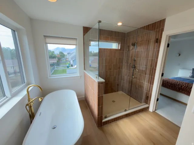 Freestanding soaking tub with a gold-finish floor-mounted faucet, glass-enclosed shower with vertical tile, and wood-finish flooring