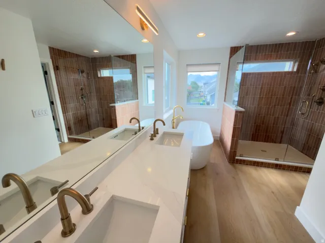 Dual vanity with undermount sinks and brass-finish fixtures, quartz countertop, freestanding soaking tub, wood-finish flooring, and a glass-enclosed shower with vertical-set tile