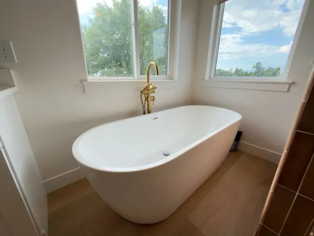 Freestanding oval bathtub with a brass floor-mounted faucet, wood-finish flooring, and multiple windows