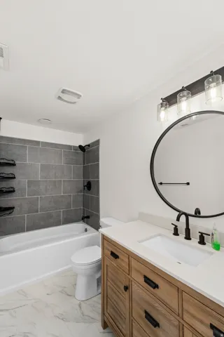 Contemporary bathroom featuring a wood-finish vanity with ample storage, a white countertop, a circular mirror, and a tub with gray tile surround