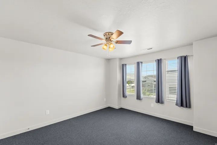 Three windows with blinds, ceiling fan with light fixture, and dark carpet flooring