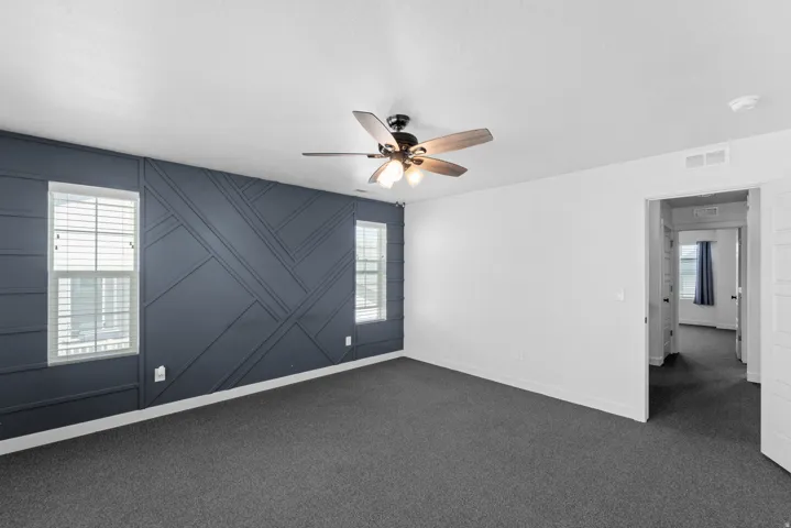 Textured accent wall with geometric paneling, dual windows with blinds, ceiling fan with integrated lighting, and neutral carpet flooring