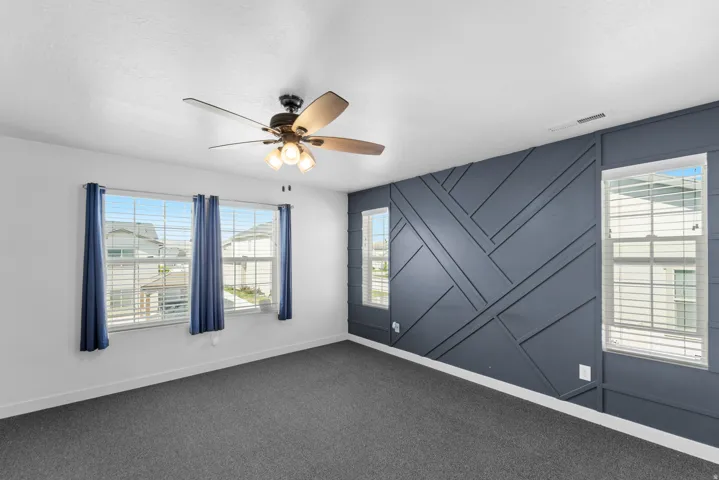 Wall-to-wall carpeting with a decorative accent wall, featuring multiple windows with blinds, and a ceiling fan with integrated lighting