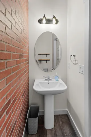 Brick accent wall, pedestal sink, oval mirror, wall-mounted light fixture, wood-finish flooring