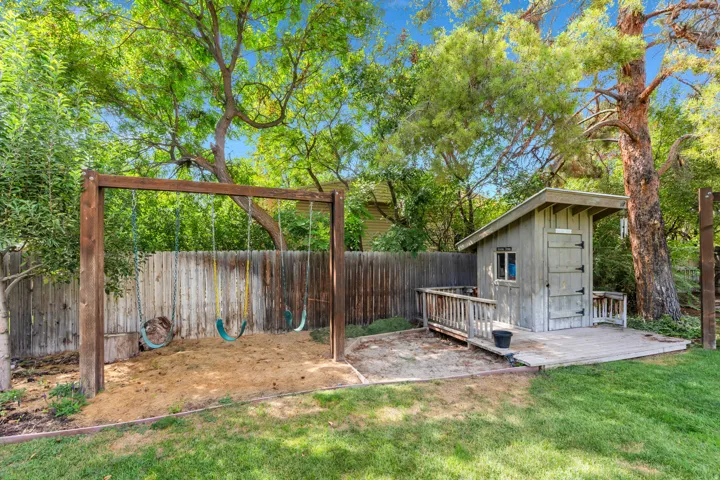 Expansive backyard featuring a manicured lawn, established trees, a wood-fenced perimeter, a swing set, and a detached shed with an attached ramp
