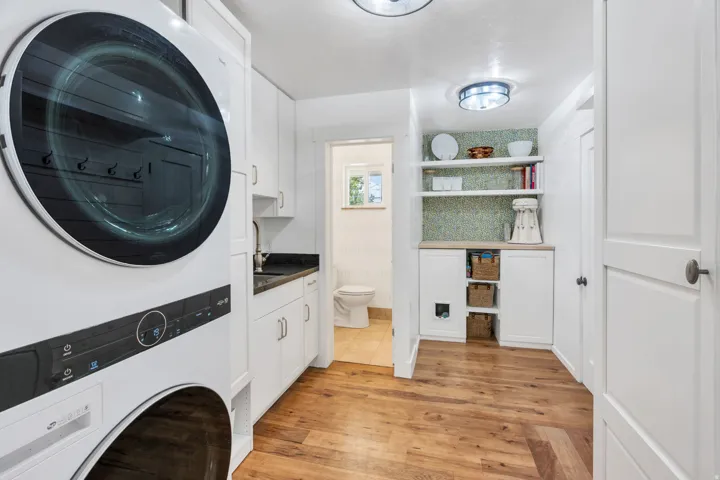 Stacked laundry appliances, wood-finish flooring, white cabinetry, dark stone countertops, and open shelving