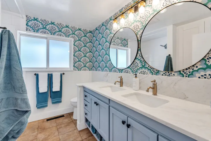 Dual round mirrors, a double vanity with light blue cabinetry, gold-finish faucets, a white solid-surface countertop with integrated sinks, and patterned wallpaper