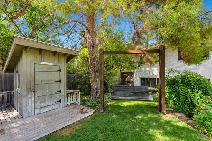 Backyard featuring a wooden storage shed, wood-plank decking, a freestanding wooden swing, mature trees, and landscaping