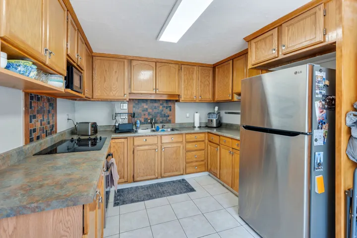 Kitchen featuring wood cabinetry, stainless steel refrigerator, integrated cooktop, tiled backsplash, and white floor tiling