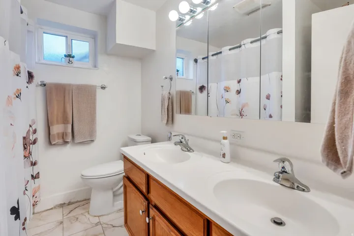 Dual white ceramic sinks with chrome faucets, wood-finish vanity with cabinet storage, mirrored medicine cabinet with integrated lighting, tiled flooring, and a white toilet