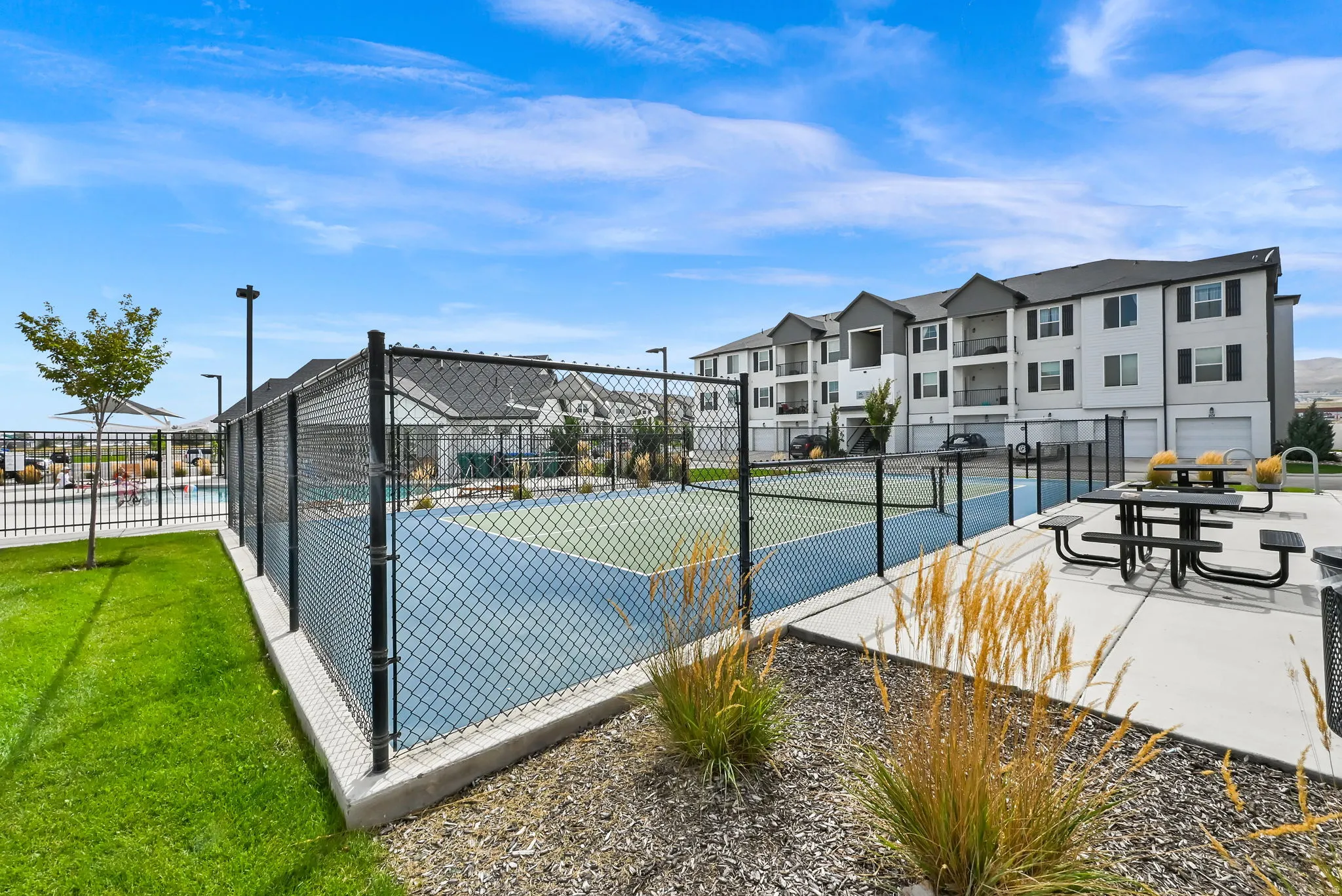 Hard court tennis court with chain-link fencing, adjacent recreational pool, and nearby outdoor dining area