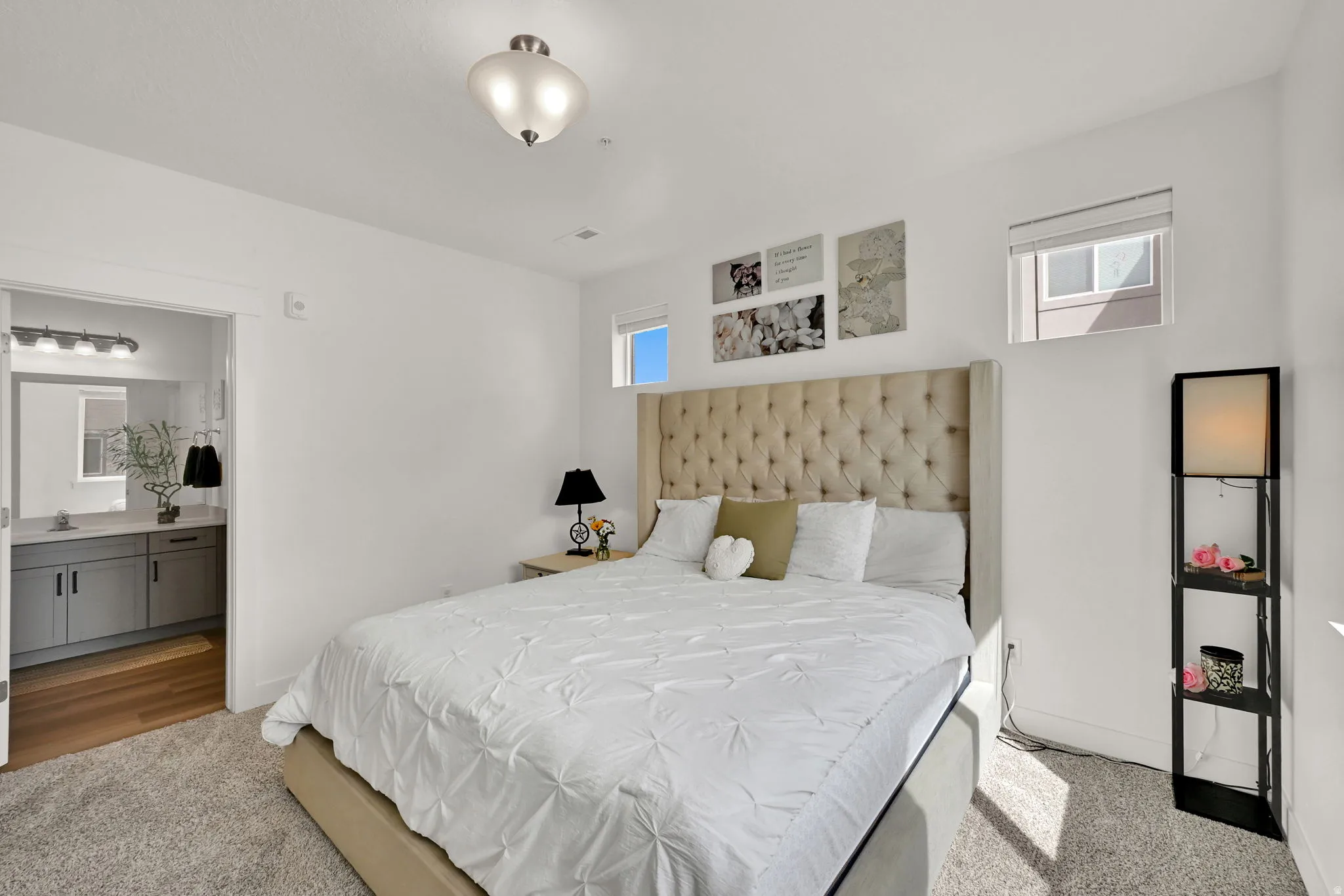 Tufted fabric headboard, recessed window, wall-mounted light fixture, vanity with dual-door cabinetry, and wood-finish flooring in the adjoining space