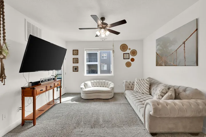 Carpeted living area featuring a ceiling fan with integrated lighting, a wall-mounted television, and a window with natural light