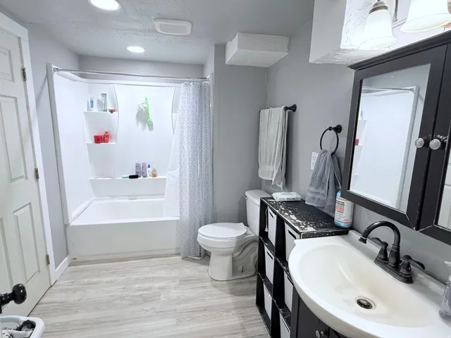 White tub and shower combination with an integrated shelving system, light wood-finish flooring, dark wood-finish vanity with an integrated sink, and a matching framed medicine cabinet