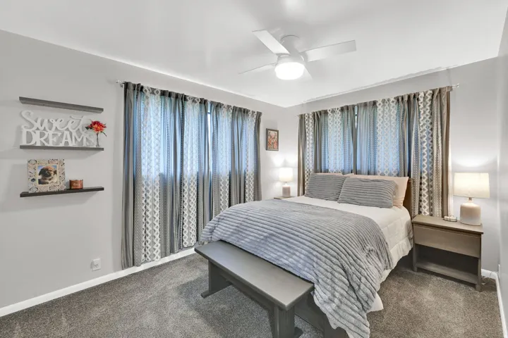 Neutral wall paint, ceiling fan with integrated lighting, carpeted flooring, floating wall shelves, and multiple windows