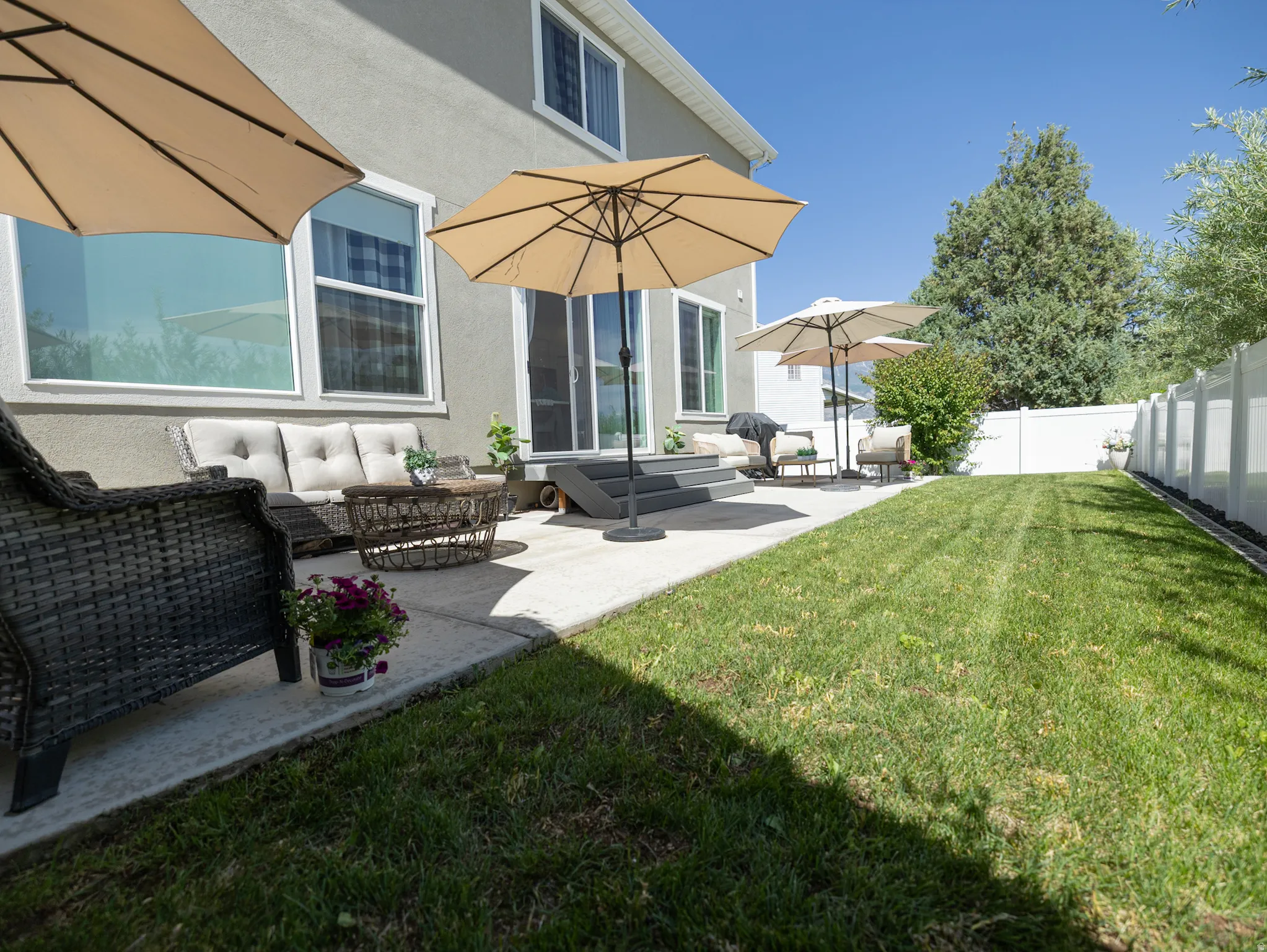 Expansive backyard featuring a concrete patio, a manicured lawn, a white privacy fence, and mature landscaping