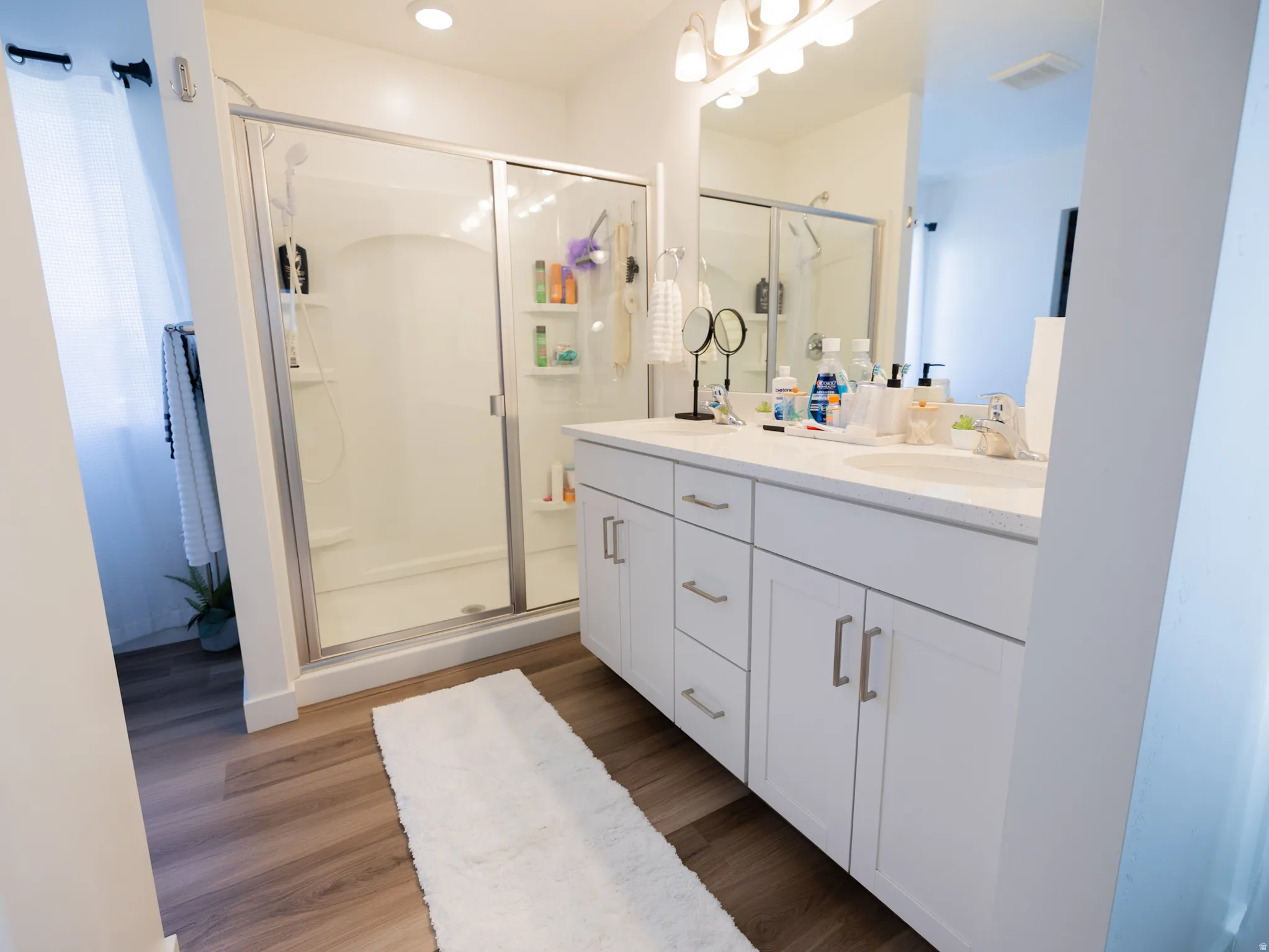 Dual vanity with white cabinetry, wood-finish flooring, recessed lighting, and a glass-enclosed shower