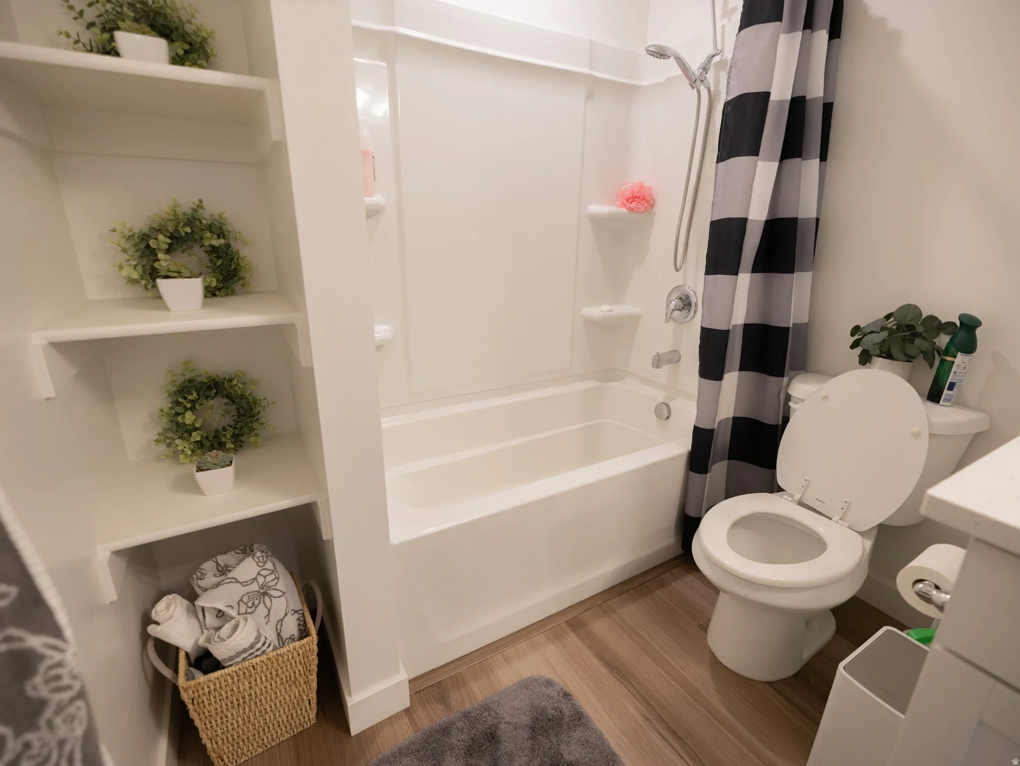 White built-in shelving, white bathtub with integrated shower, chrome fixtures, white toilet, and wood-finish flooring