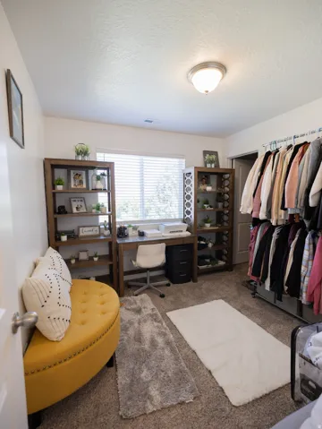 Built-in desk with shelving, window with horizontal blinds, overhead flush mount lighting, and carpeted flooring