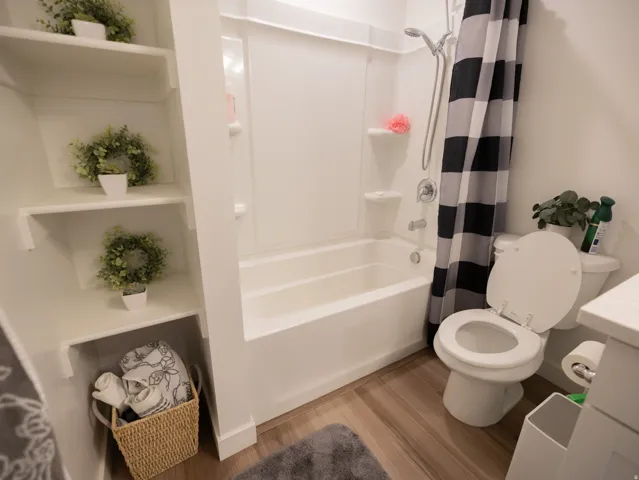 White built-in shelving, white bathtub with integrated shower, chrome fixtures, white toilet, and wood-finish flooring