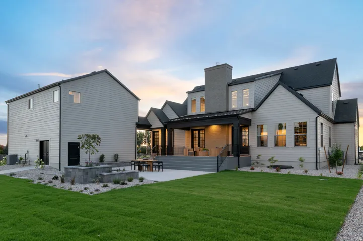 Exterior featuring light gray siding, dark-framed windows, a covered patio with recessed lighting, a stone-clad chimney, and a meticulously landscaped yard with a concrete patio