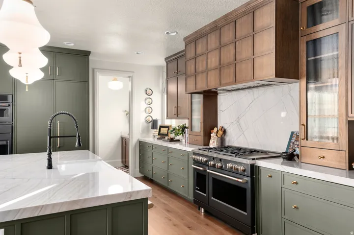 Kitchen with green cabinetry, white stone countertops, a large center island with an integrated sink, a professional-grade range, and wood-finish upper cabinetry with reeded glass accents