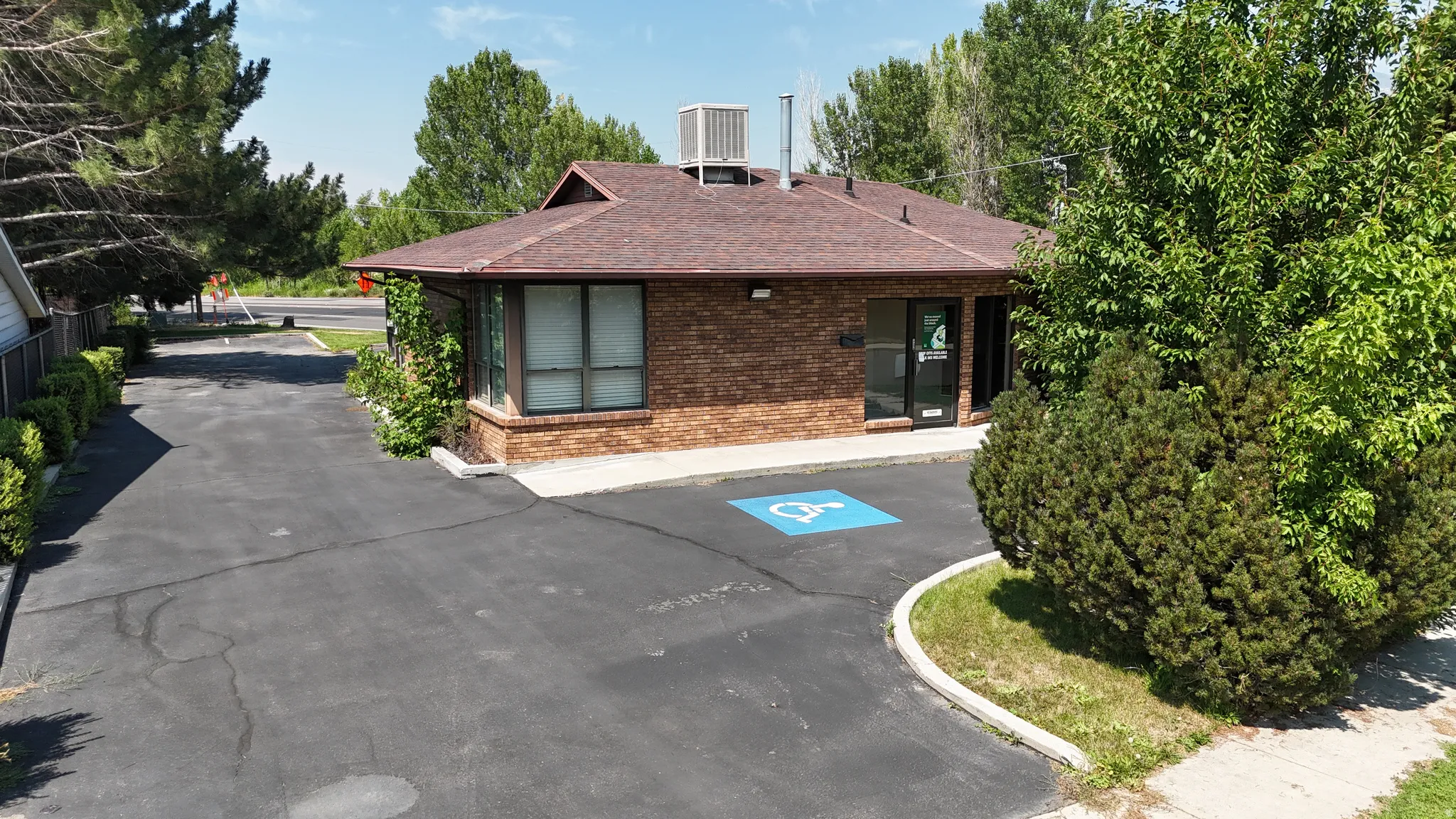 Brick exterior with a brown shingle roof, paved parking area with designated accessible space, and mature perimeter landscaping